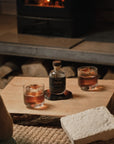 Two glasses of whiskey and a bottle labeled 'Sylva' on a wooden table with a et of 2 NUDE Big Top DOF glasses and a fireplace in the background.