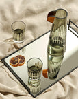 A clear crystal carafe and two tumblers with a geometric pattern, displayed on a reflective surface, accompanied by dried orange slices and a beige textile backdrop.