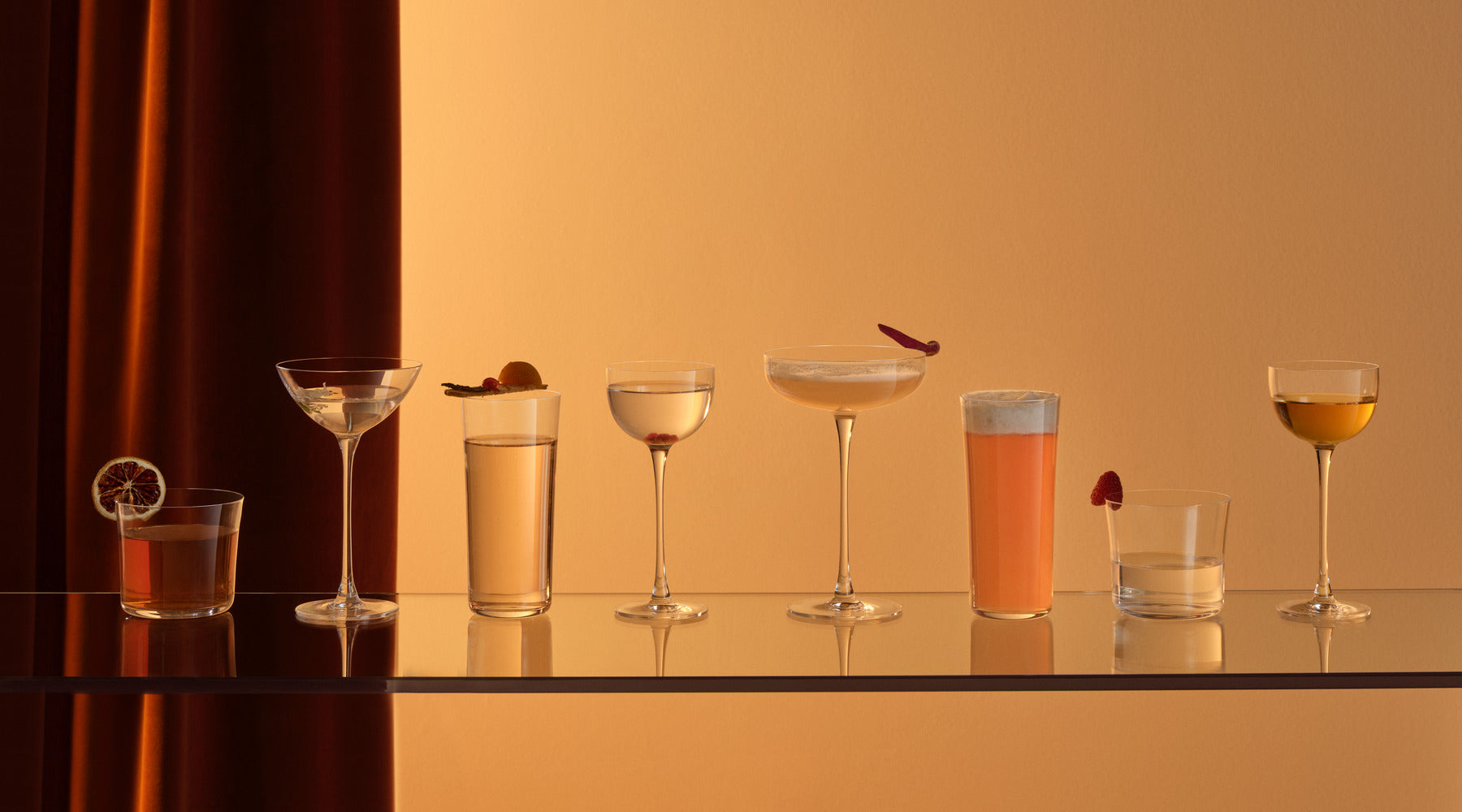 NUDE Savag cocktail collection, a coupe glass, coupetini glass, pony glass, highball, water and lowball glass presented in an orange scenery and orange toned cocktails