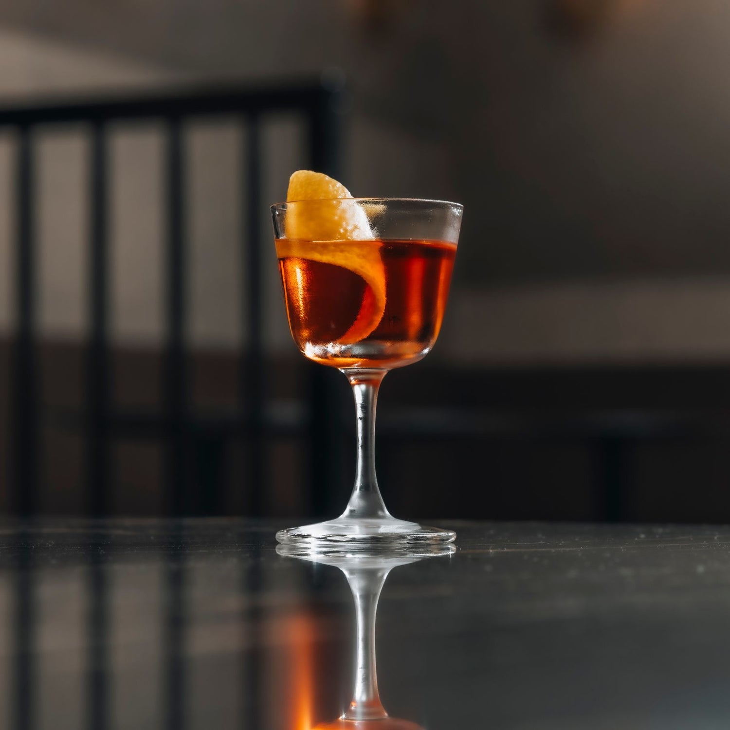 A NUDE BarGiani welcome drink glass filled with an amber-orange cocktail and garnished with a curled orange peel, set on a dark reflective surface with a blurred indoor background.