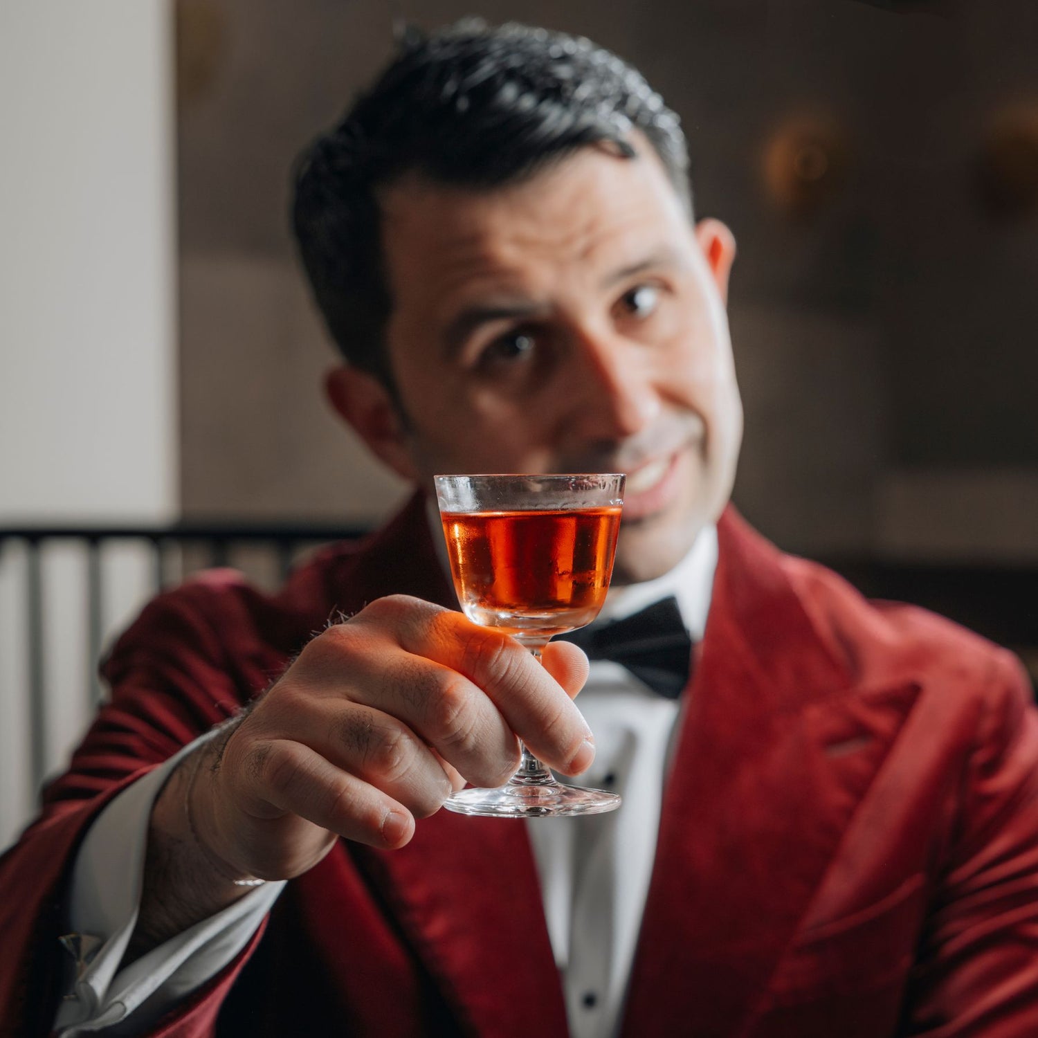 Giorgio BarGiani in a red velvet blazer, white shirt, and black bow tie holds up the NUDE Welcome drink glass with amber liquid, likely a whiskey or liqueur, in a dimly lit indoor setting.