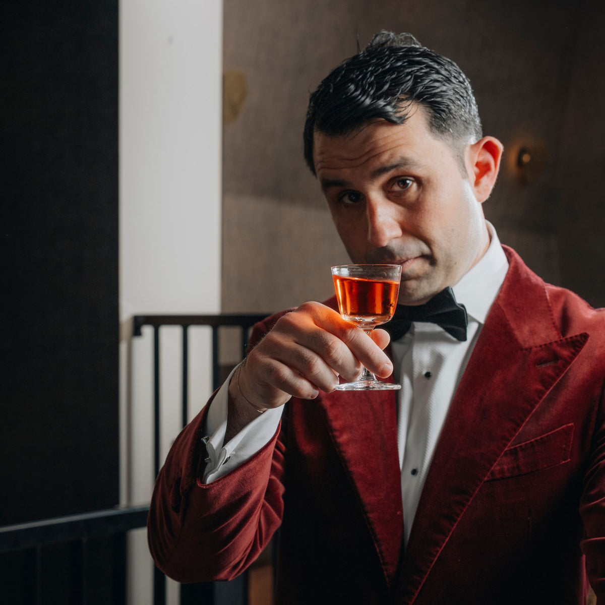 Giorgio Bargiani in a red velvet tuxedo jacket, white shirt, and black bow tie holds up his NUDE welcome drink glass with amber liquid, looking directly at the camera in an indoor setting.