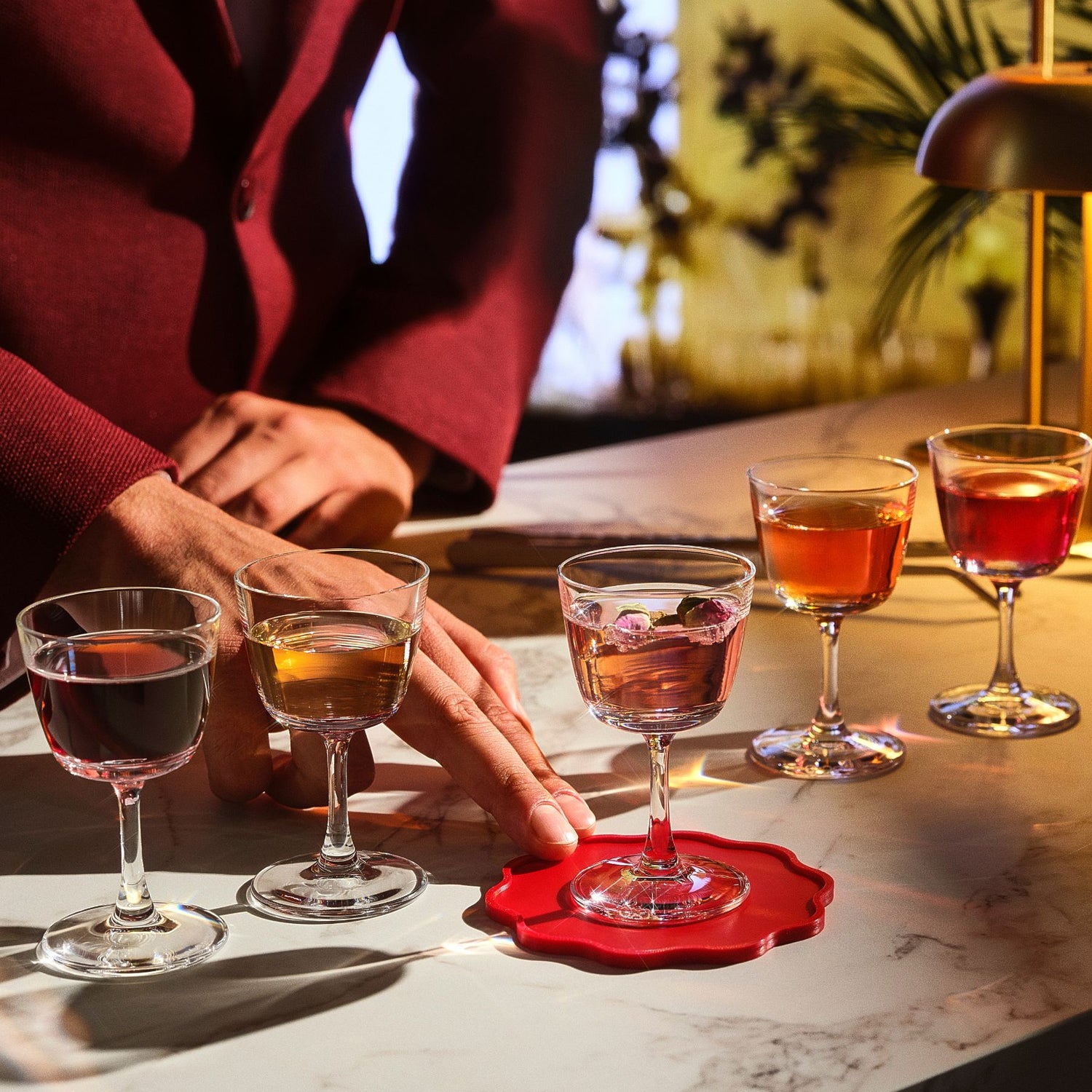A close-up lifestyle shot of a person in a burgundy blazer placing a pink cocktail on a scalloped red coaster on a white marble bar counter. Four other NUDE Bargiani welcome drink glasses are lined up behind it on a warm, dimly lit bar table.
