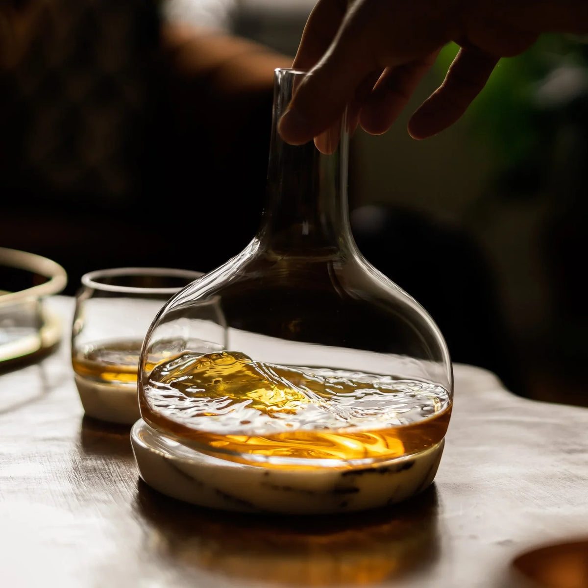 NUDE Glass Chill whiskey decanter with whiskey on a wooden table, hand reaching towards it.
