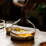 NUDE Glass Chill whiskey decanter with whiskey on a wooden table, hand reaching towards it.