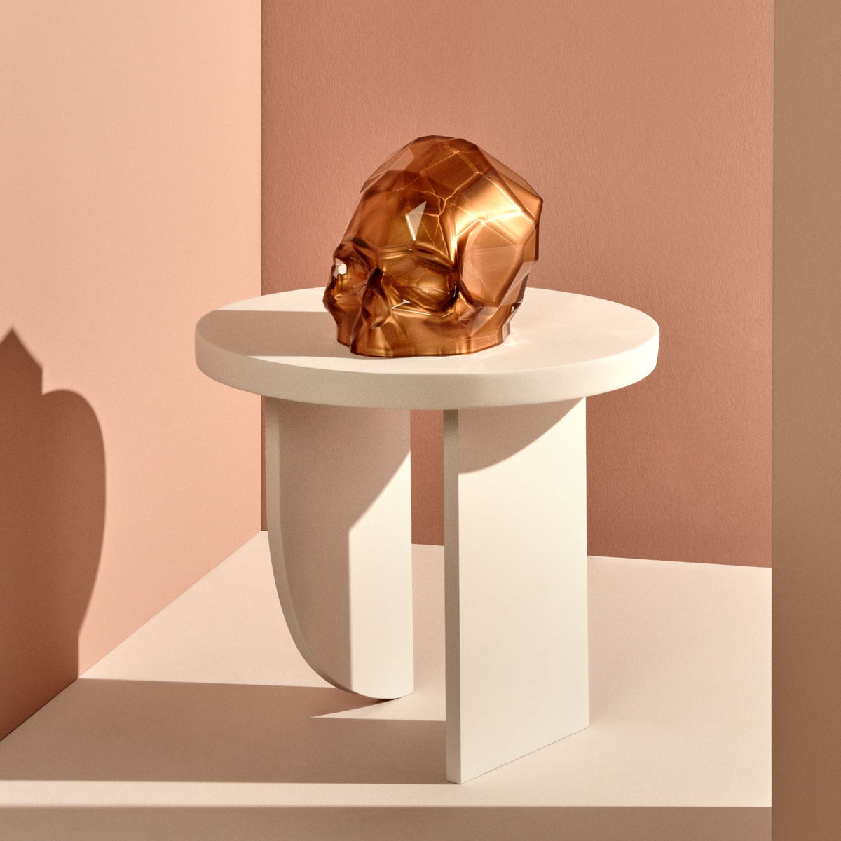 The Memento Mori Faceted Skull in copper on a round white sculptural side table against a dusty rose wall, with a dramatic cast shadow.