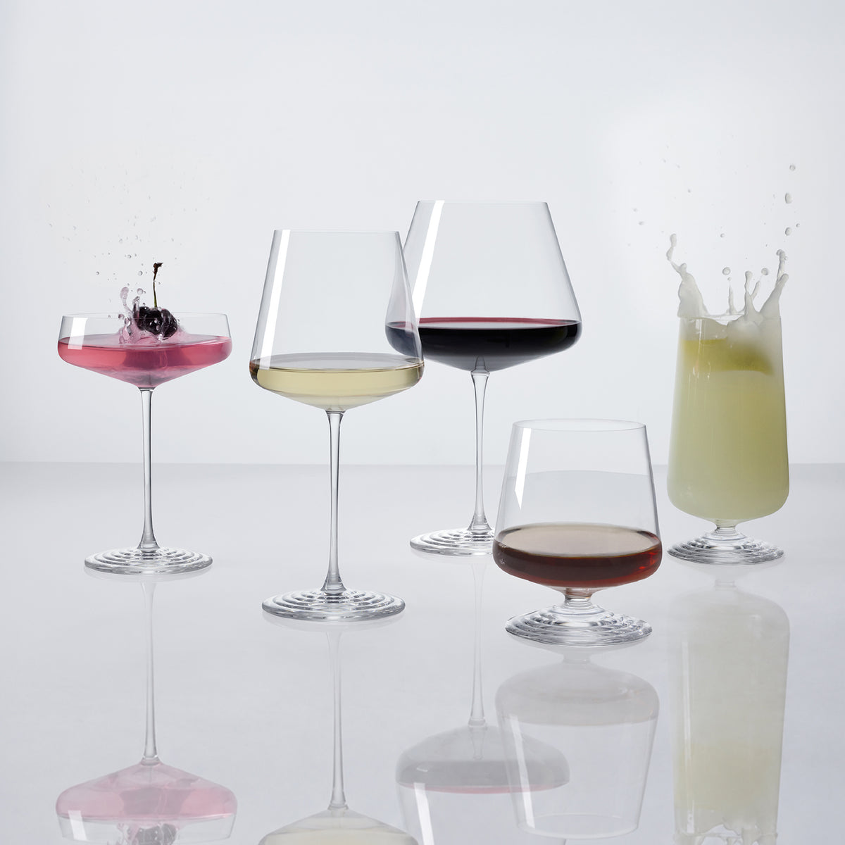 The five NUDE | Tom Dixon Splash glasses in a row, each filled: coupe with pink cocktail and cherry garnish splashing, white wine, red wine, lowball with amber spirit, and highball with a pale green drink splashing, all on a white reflective surface