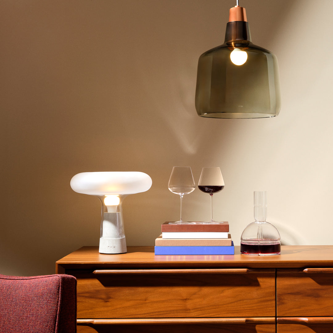 A modern table lamp with a white mushroom-shaped shade and a clear cylindrical base sits on a wooden sideboard, lit and casting a warm glow. Above it, a dark green glass pendant light hangs in a softly lit interior setting with wine glasses, books, and a decanter on the cabinet.