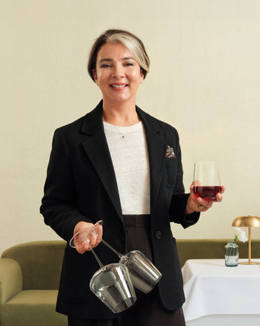 Sabiha Apaydin in a black blazer and white top holds NUDE Sera red and all purpose glass in one hand and the tumbler in the other, standing in an indoor setting with a table and lamp in the background.