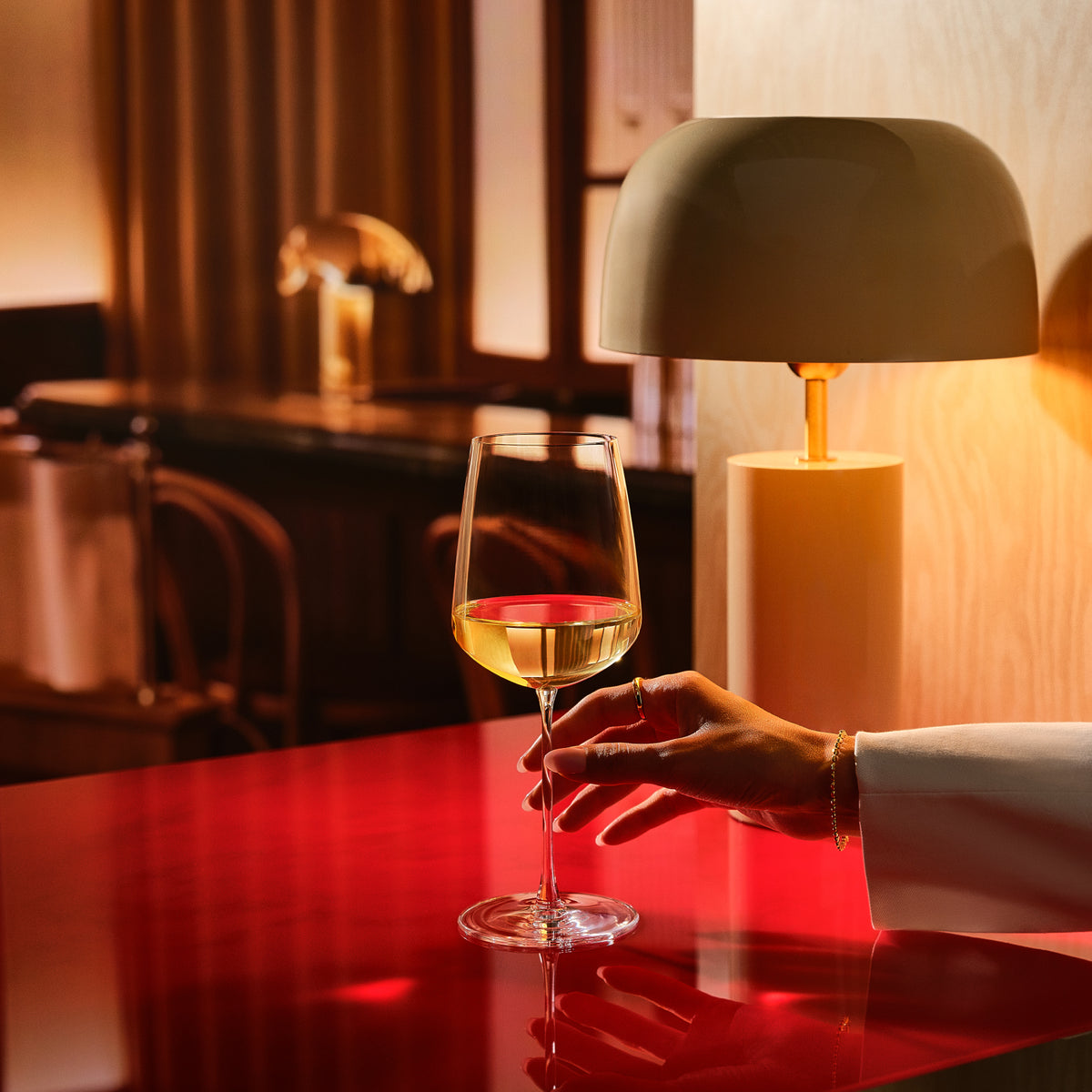 A hand in a white sleeve holds a Sera All purpose wine glass with a small amount of white wine on a glossy red table, with a warm lamp and dark interior softly blurred in the background.