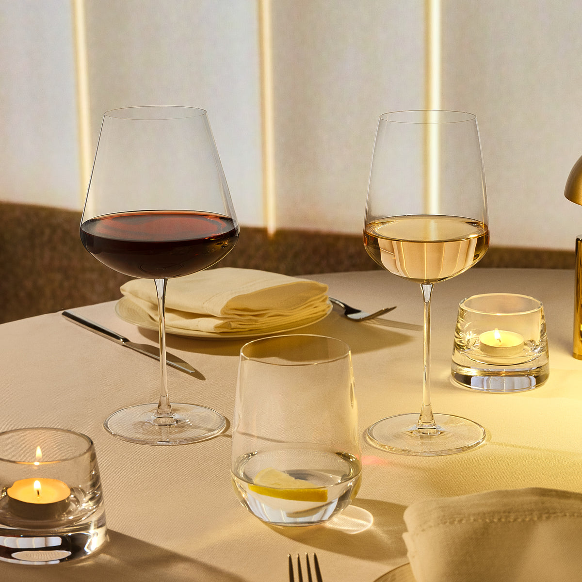 Two clear NUDE Sera wine glasses on a softly lit dining table, one filled with red wine and one with white wine, alongside candle holders, folded napkins, and cutlery.