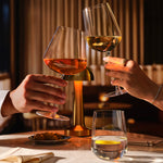Two hands holding NUDE Sera wine glasses in a warmly lit restaurant setting, with one glass containing red wine and the other containing white wine, clinking together above a table with a small plate of olives and a glass of water in the foreground.