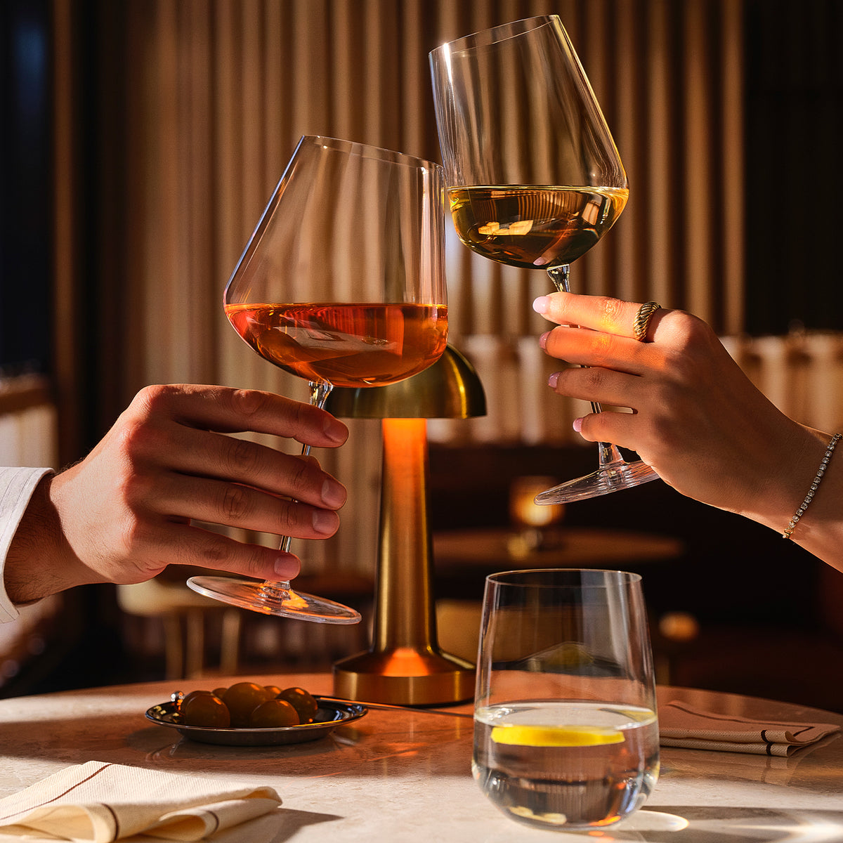 Two hands holding NUDE Sera wine glasses in a warmly lit restaurant setting, with one glass containing red wine and the other containing white wine, clinking together above a table with a small plate of olives and a glass of water in the foreground.