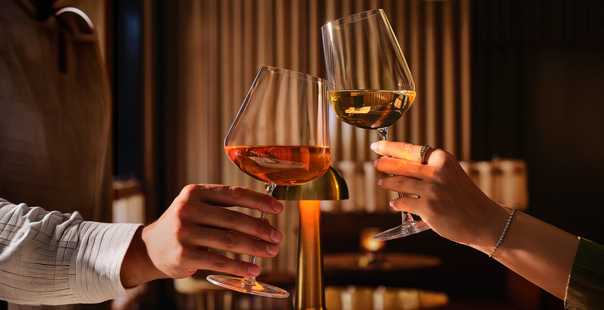 Two people clinking  NUDE Sera wine glasses in a warmly lit indoor setting. One glass contains a pinkish-red wine, and the other contains a pale yellow wine.