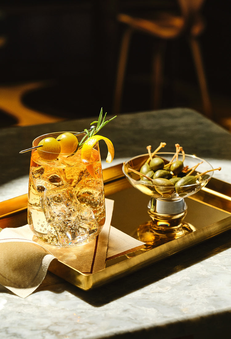 A tumbler of teh NUDE Sera collection filled with a clear amber drink, ice, a green olive, and a curled orange peel garnish, placed on a gold tray beside a small glass bowl of olives in a dimly lit bar-like setting.