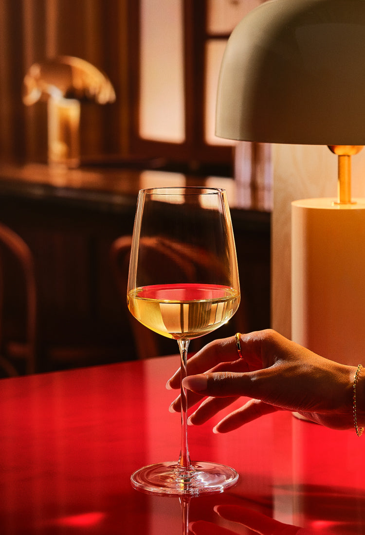 All Purpose wine glass of the NUDE Sera collection containing white wine sits on a glossy red table, held by a person's hand in a warmly lit indoor setting with a lamp in the background.