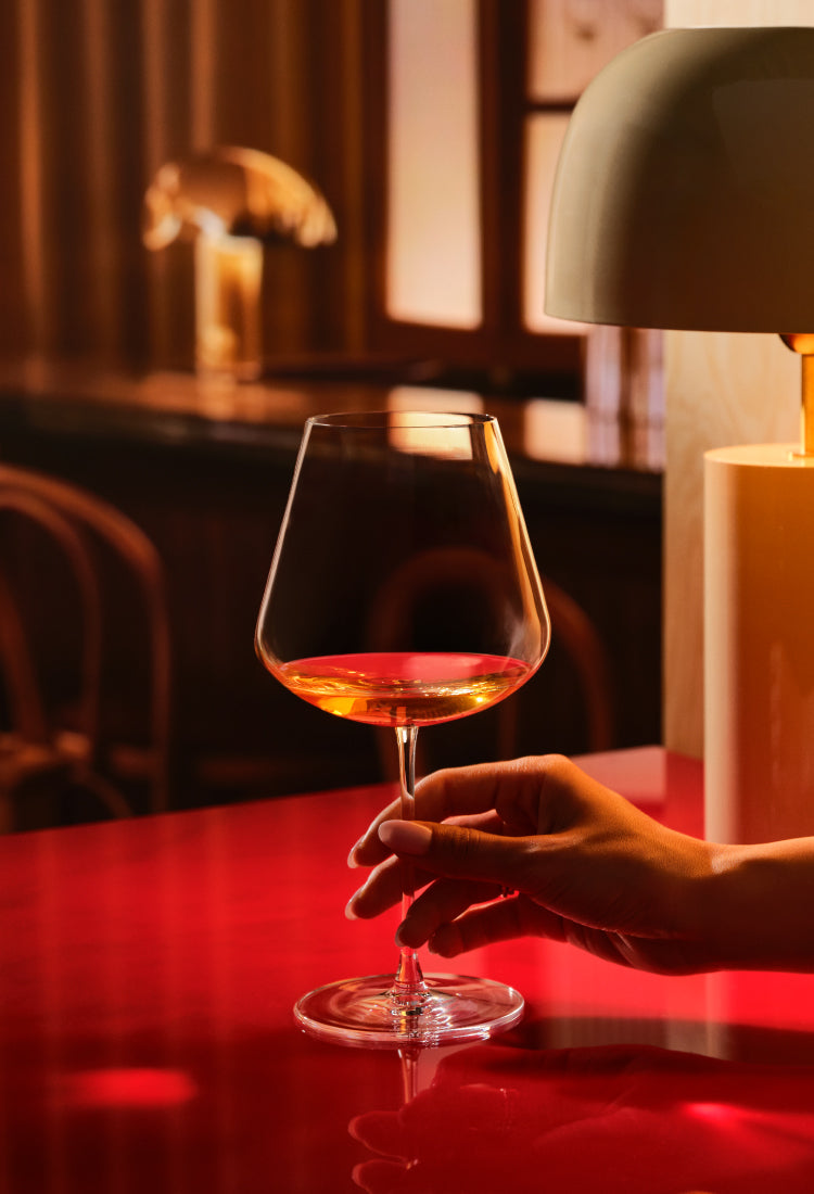 The Red Burgundy wine glass of the NUDE Sera collection with a small amount of amber liquid, held by a hand over a glossy red table in a warmly lit interior setting with a lamp in the background.