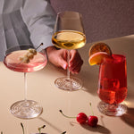 Three clear stemmed drinking glasses of the NUDE x Tom Dixon Splash collection arranged on a beige tabletop with drinks and a person's hand and torso partially visible behind them. The left glass holds a pink cocktail with a cherry garnish, the center glass holds a pale yellow wine, and the right glass holds a red cocktail with a citrus slice and cherry garnish.