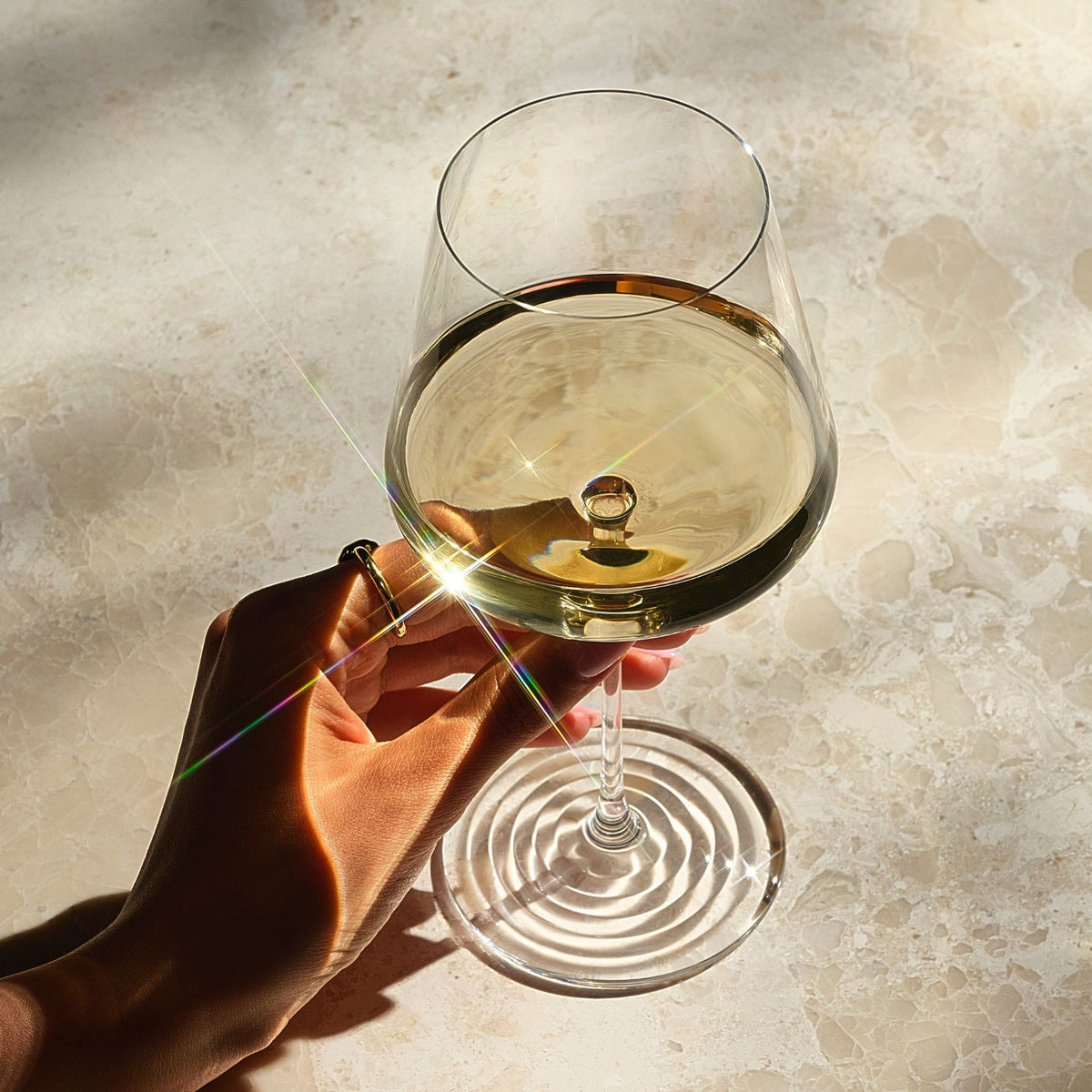 A hand holds a NUDE glass Splash clear wine glass containing pale golden liquid on a sunlit beige stone surface, with strong shadows and lens flare across the scene.