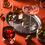 Three NUDE Splash glasses styled on a warm terracotta backdrop: a lowball with amber spirit and ice on a silver tray, a coupe with pink cocktail splashing and edible flower garnish, and a lowball with dark red drink and ice in the foreground, surrounded by olives, cocktail picks, and cinnamon sticks.