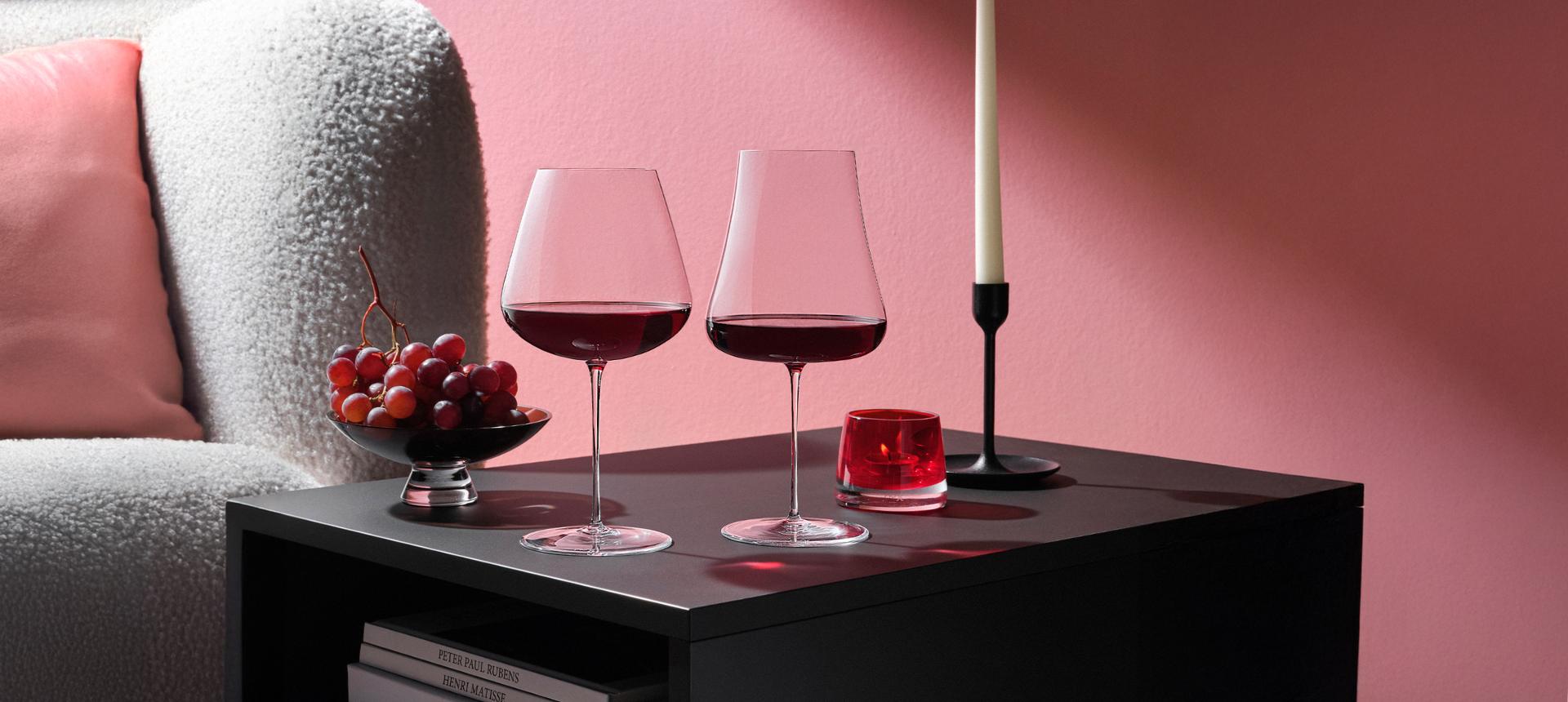 NUDE Stem Zero Volcano Red Wine Glass and Elegant Red Wine Glass, styled on a black side table with grapes, candle, and pink boucle sofa.