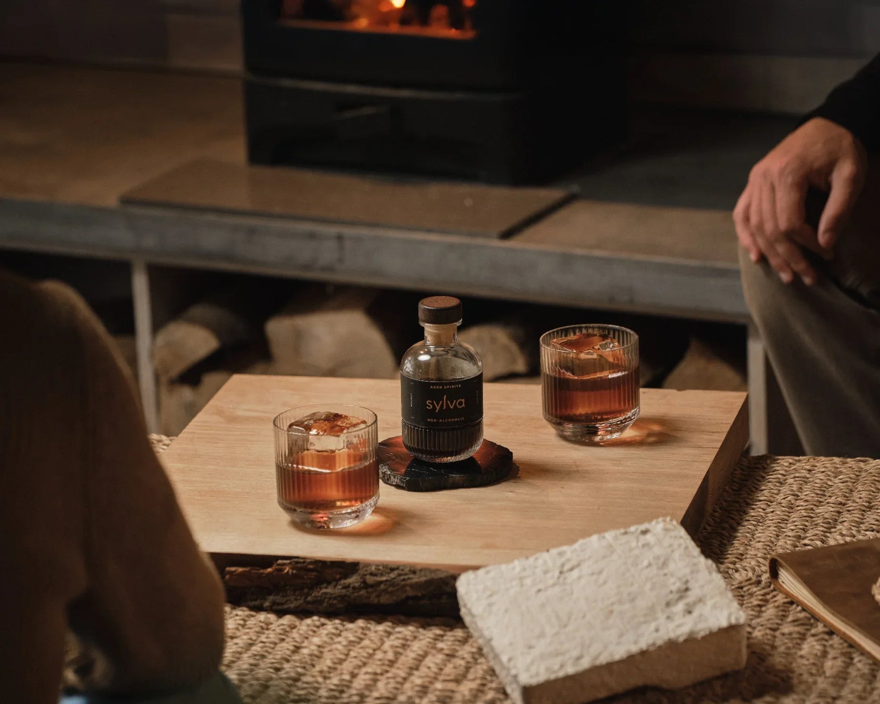 Two glasses of whiskey and a bottle labeled 'Sylva' on a wooden table with a et of 2 NUDE Big Top DOF glasses and a fireplace in the background.