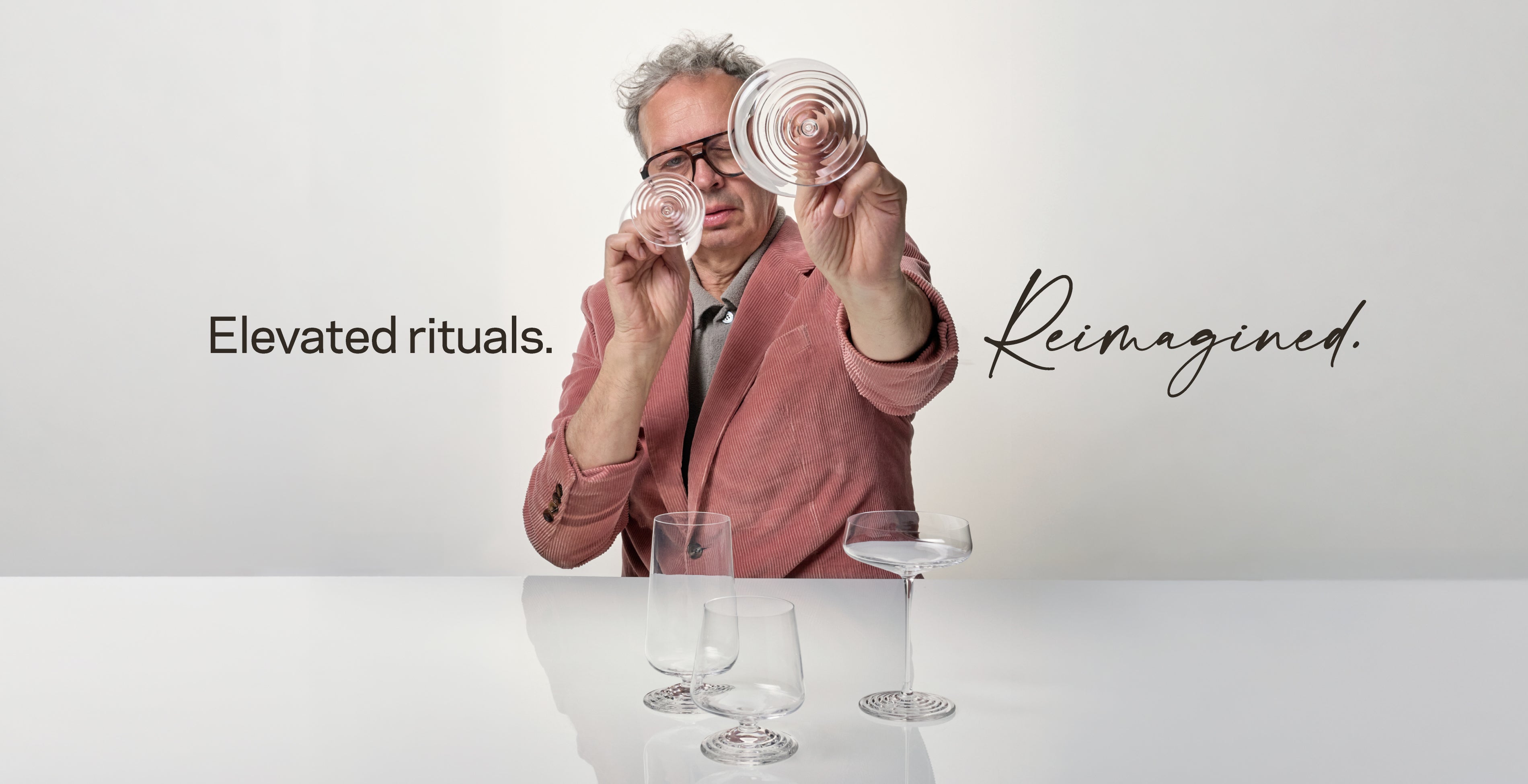 Tom Dixon in a pink blazer holds a spiral glass sculpture above a table with several clear drinking glasses of the NUDE x Tom Dixon Splash Collection, against a light gray studio background with the text 'Elevated rituals. Reimagined.'