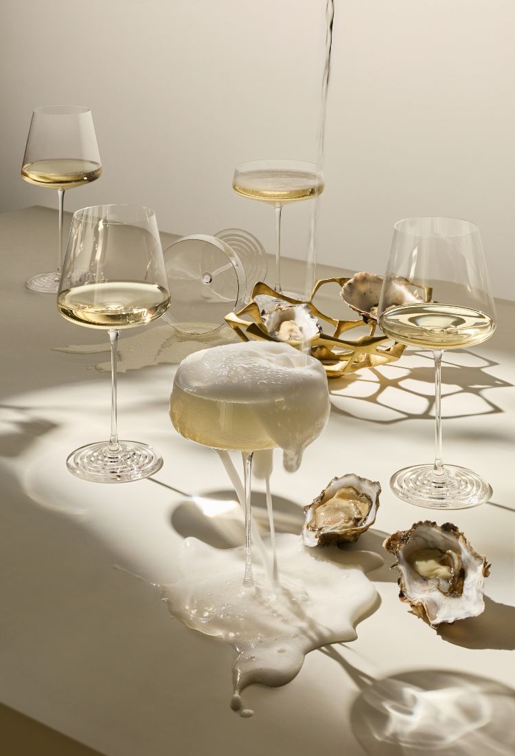 A styled still life with several clear wine glasses  of the NUDE x Tom Dixon Splash collection containing white wine, a central glass of creamy white liquid with foam, oyster shells, and a gold geometric object on a light surface under warm directional light.