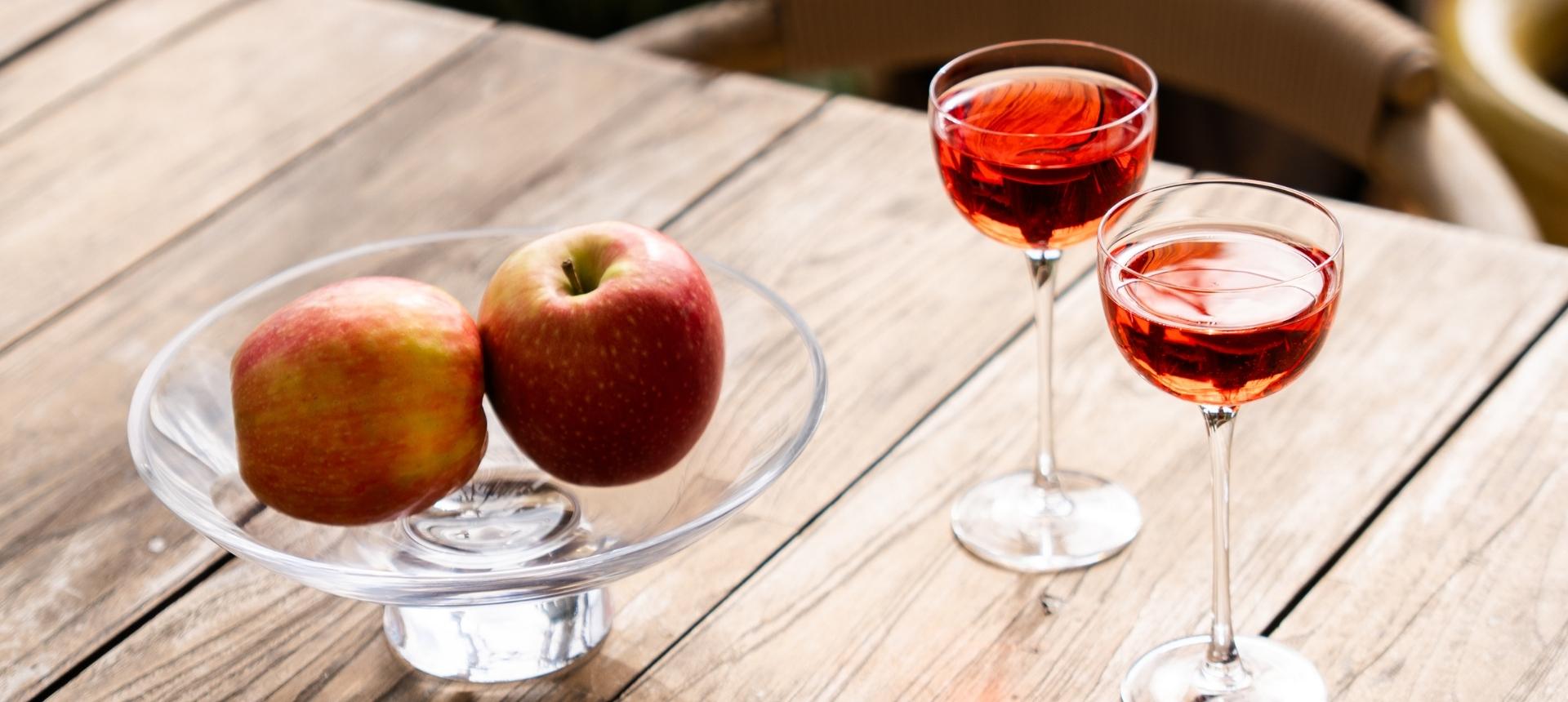 Silhouette bowl with two apples beside two Savage Pony glasses filled with rosé, on a sunlit wooden outdoor table.