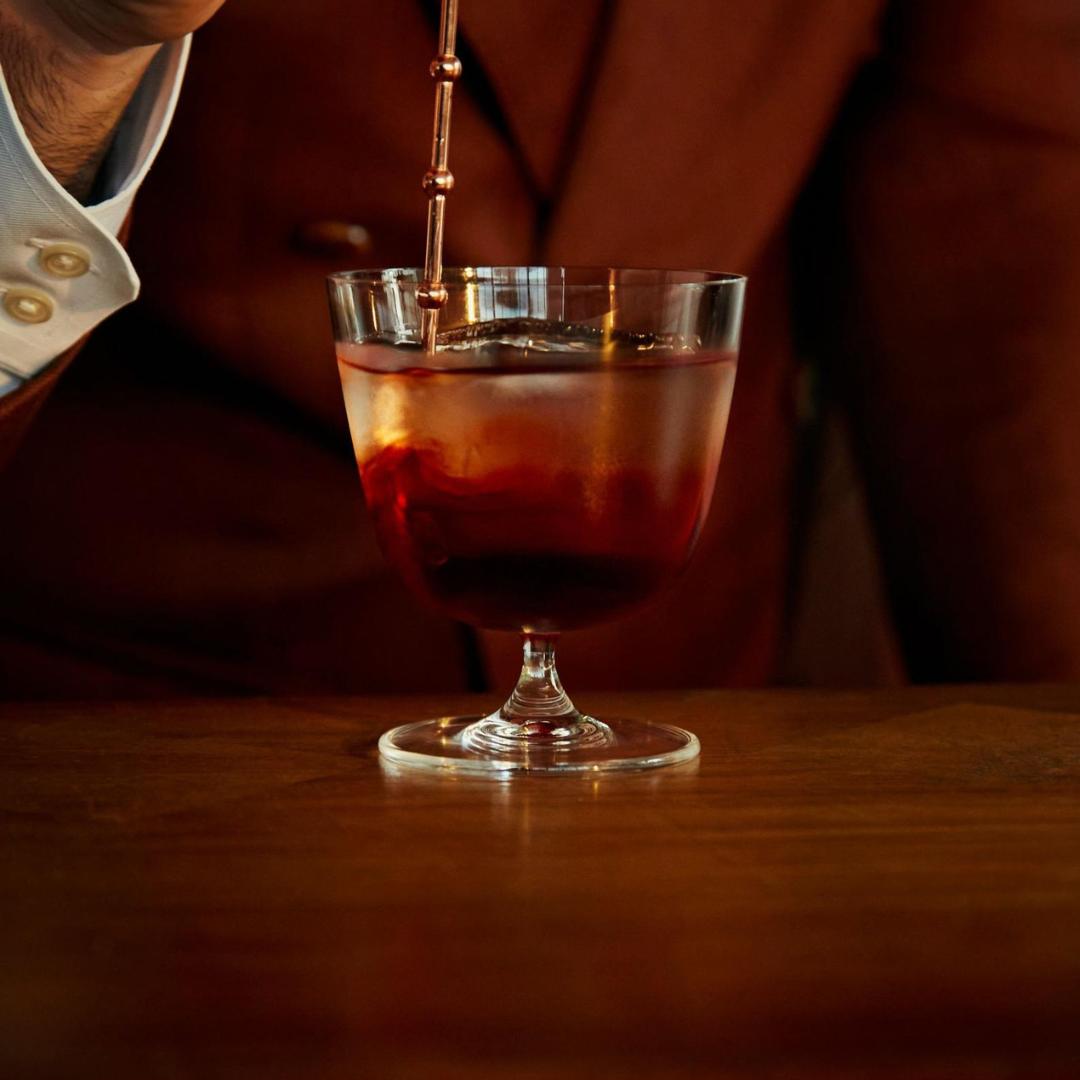 Barista pouring a dark liquid into a bar/giani cocktail glass short on a wooden surface