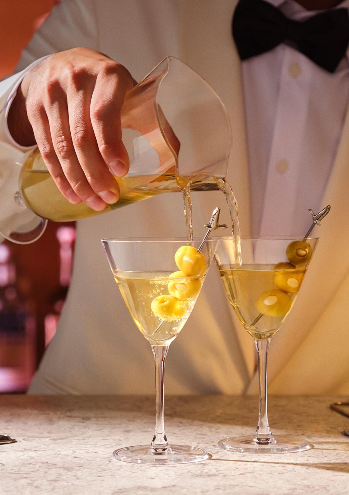 A bartender in a white dinner jacket pouring two martinis at once from the NUDE Bar/Giani Double Pour Mixing Glass.