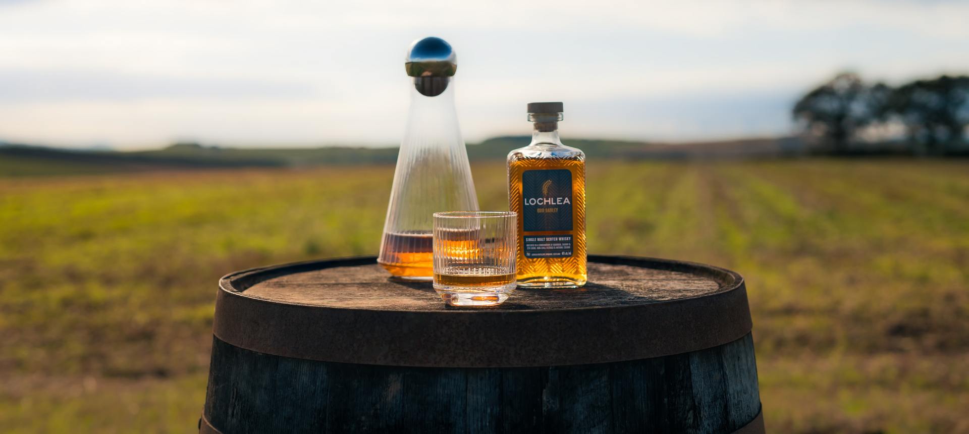 Two bottles of Lochlea whisky and a NUDE Glass Big Top whisky glass and decanter on a barrel with a scenic background