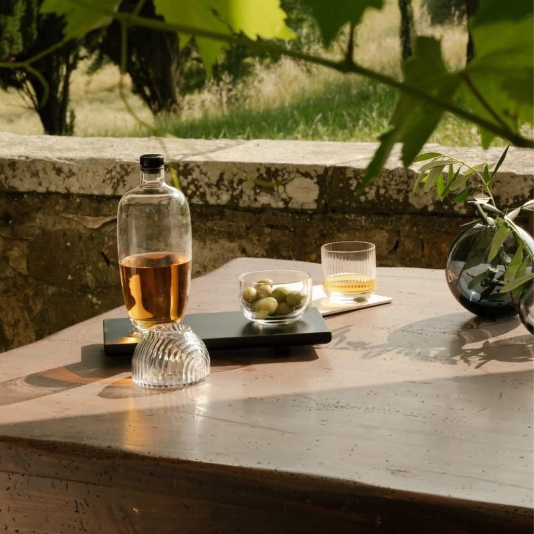 NUDE Glass Big Top Whisky Glass and Malt Decanter on a black serving tray with a Malt Bowl filled with green olives, alongside a NUDE Glass Mars Vase with olive branches, set on a stone table beneath vine leaves in warm afternoon sunlight.