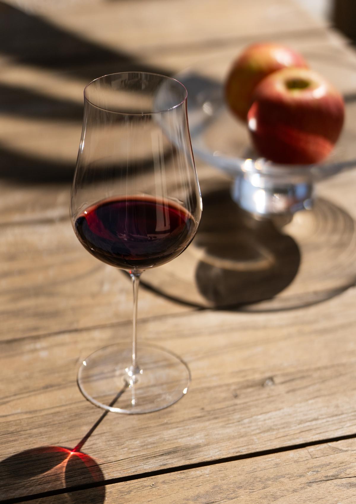NUDE Glass Ghost Zero Tulip Red Wine Glass filled with red wine, standing on a weathered wooden table in bright sunlight, with a bowl of red apples softly blurred in the background.