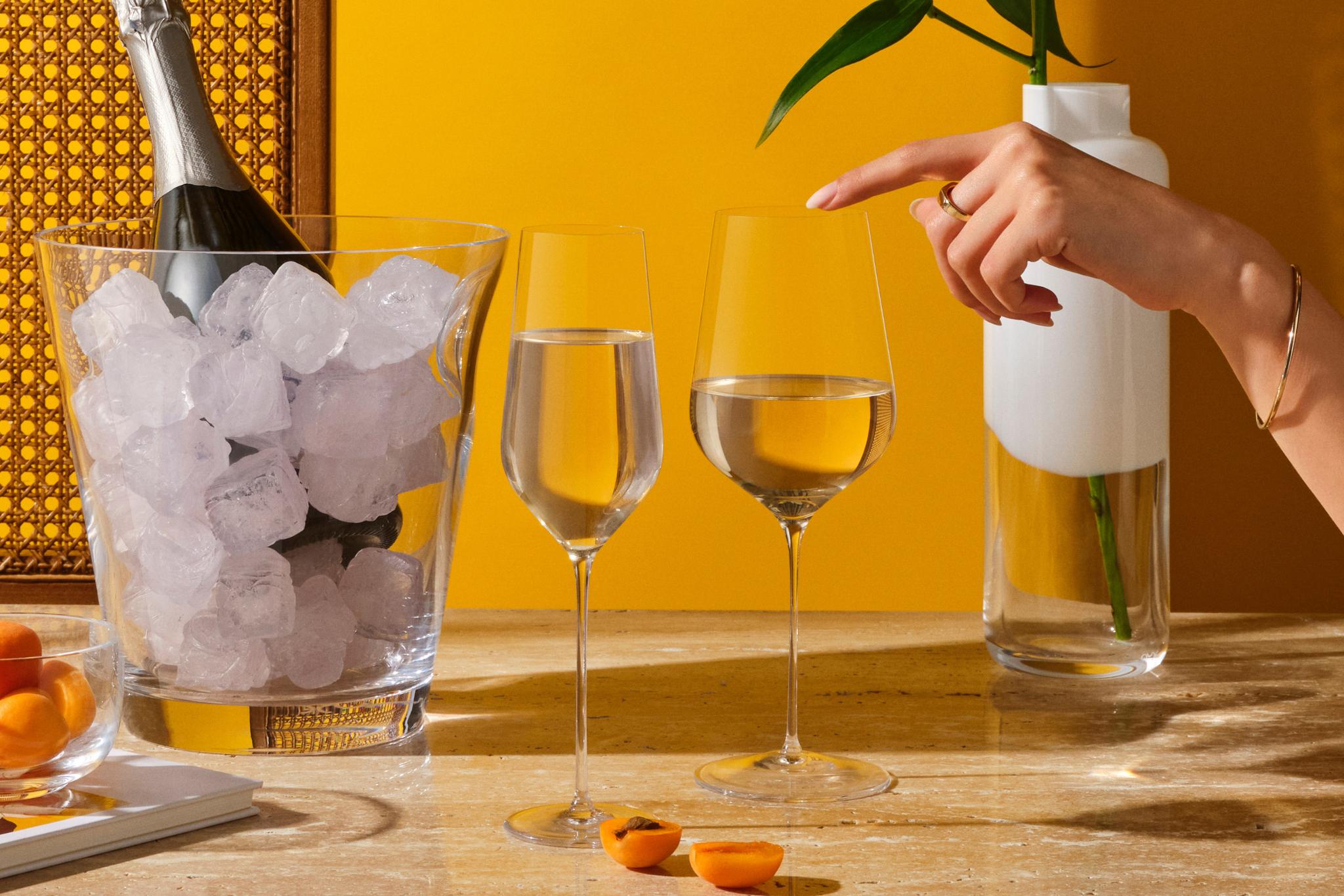 A NUDE Glacier champagne cooler holding a chilled bottle, with a NUDE Stem Zero Trio flute and white wine glass and a NUDE Magnolia vase on a sunlit table.