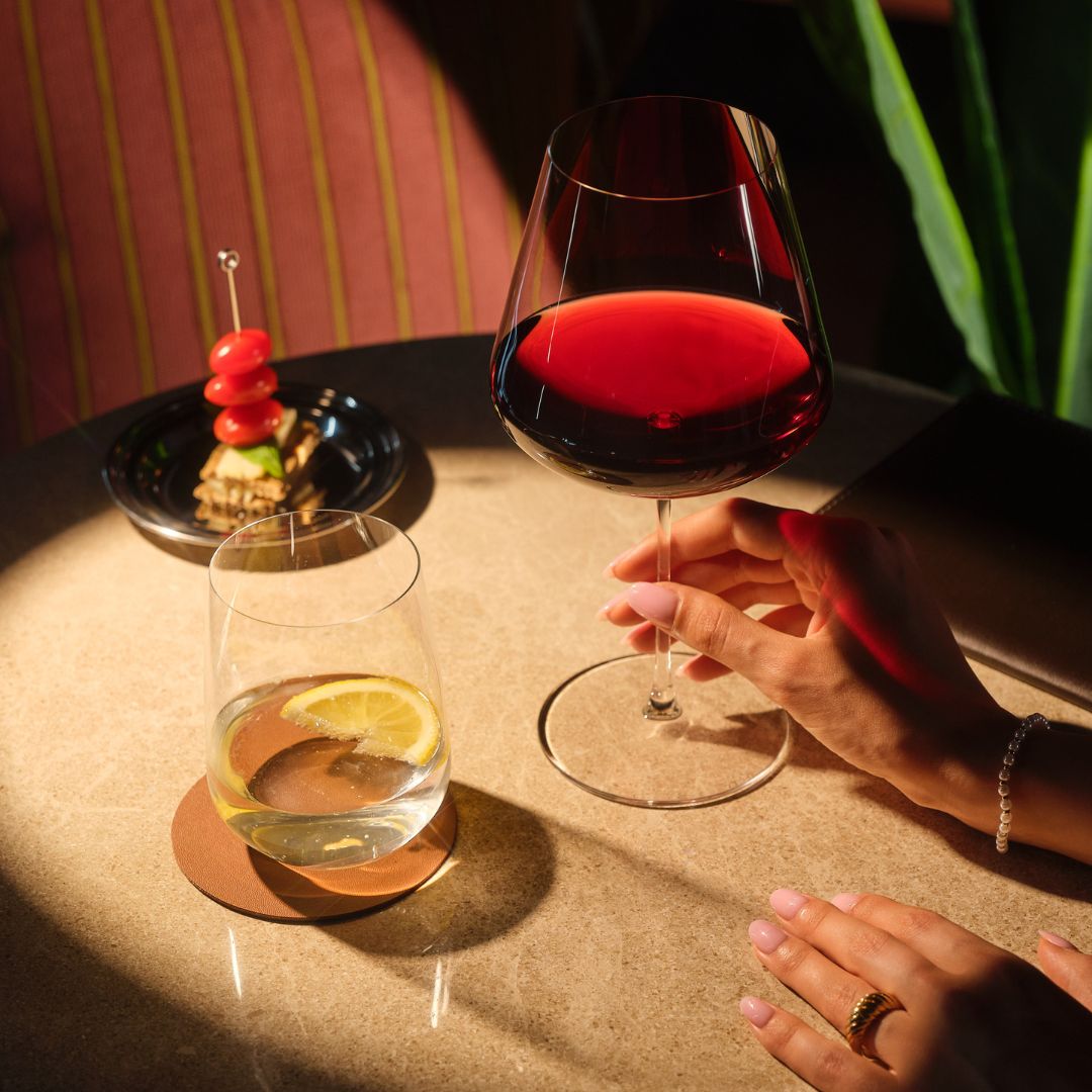 A hand holds a Sera Burgundy glass of red wine beside a Sera Tumbler of water and lemon, with a skewered appetizer on a small plate nearby