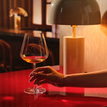 A hand holds a Sera Burgundy glass of orange wine on a glossy red table, lit by a warm table lamp in a dark wood bar setting