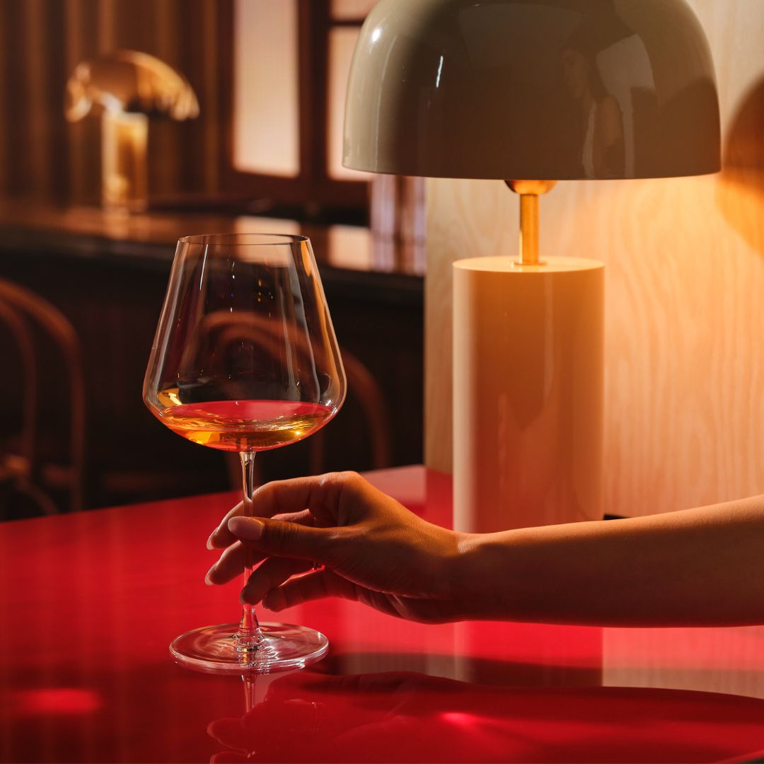 A hand holds a Sera Burgundy glass of orange wine on a glossy red table, lit by a warm table lamp in a dark wood bar setting