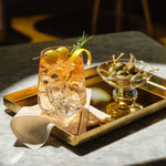 A styled cocktail in a Sera tumbler glass with ice, garnished with green olives, a lemon twist, and a rosemary sprig, served on a gold tray alongside a small glass Silhouette bowl of green olives on a stone tabletop.