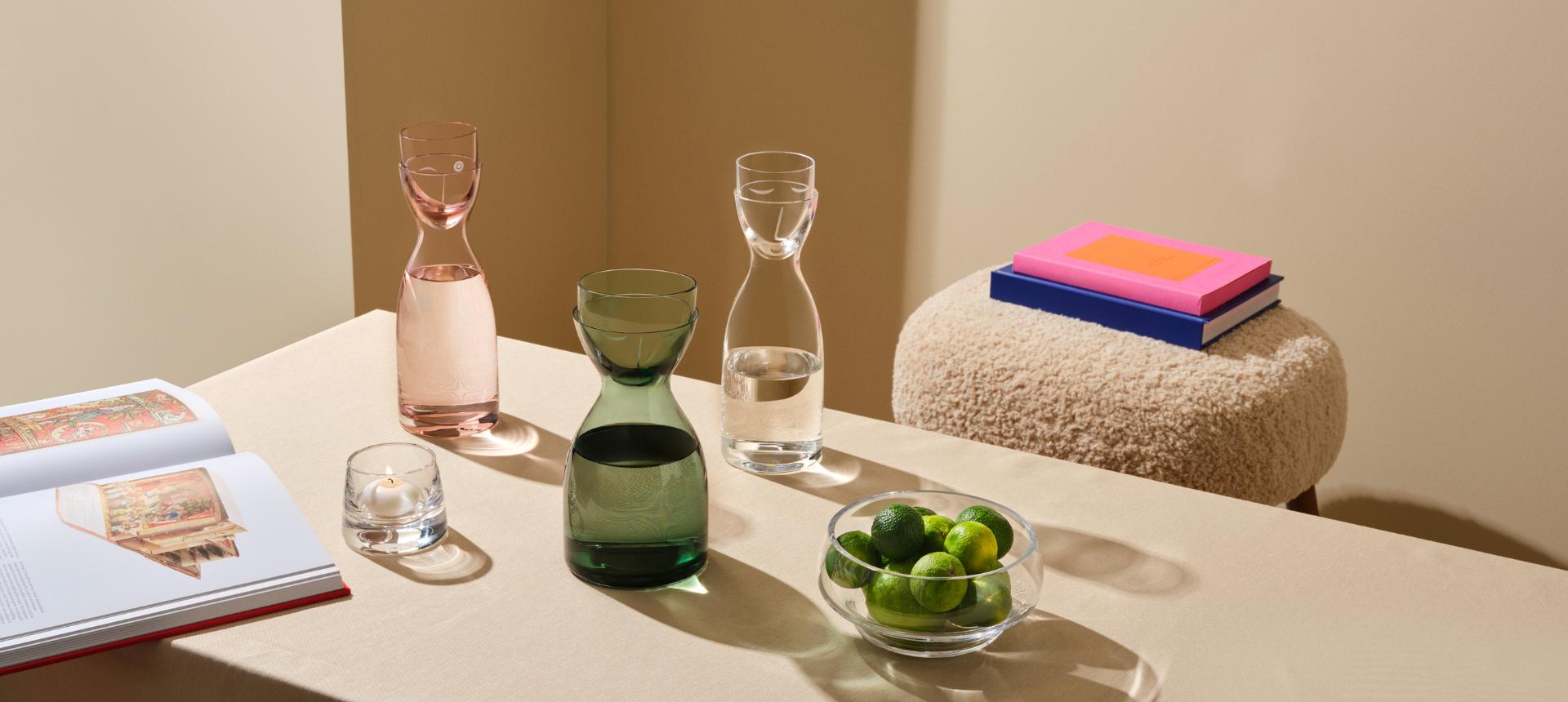 NUDE Mr & Mrs Water Set carafes in pink and green with matching tumblers, Heads Up bowl filled with limes, and Mist votive holder styled on a table with books and a boucle stool