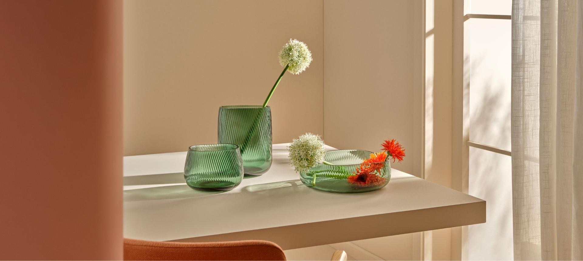 A tall and medium green NUDE Opti vase and Opti centrepiece bowl with flowers on a sunlit table.