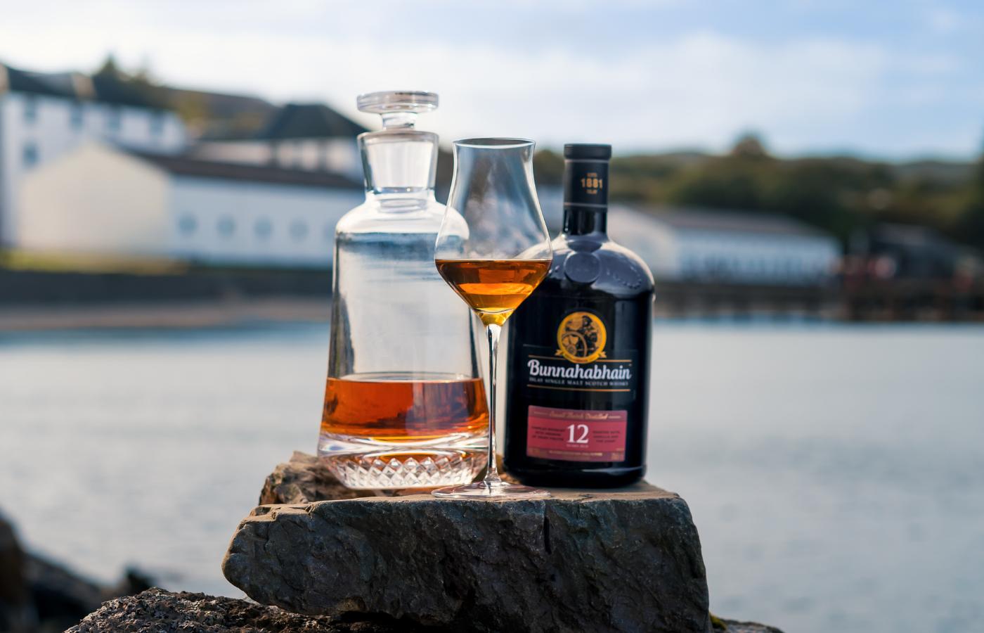 Bottle of Bunnahabhain 12-year-old whisky with a glass of Islands whisky on a stone surface by a waterfront.