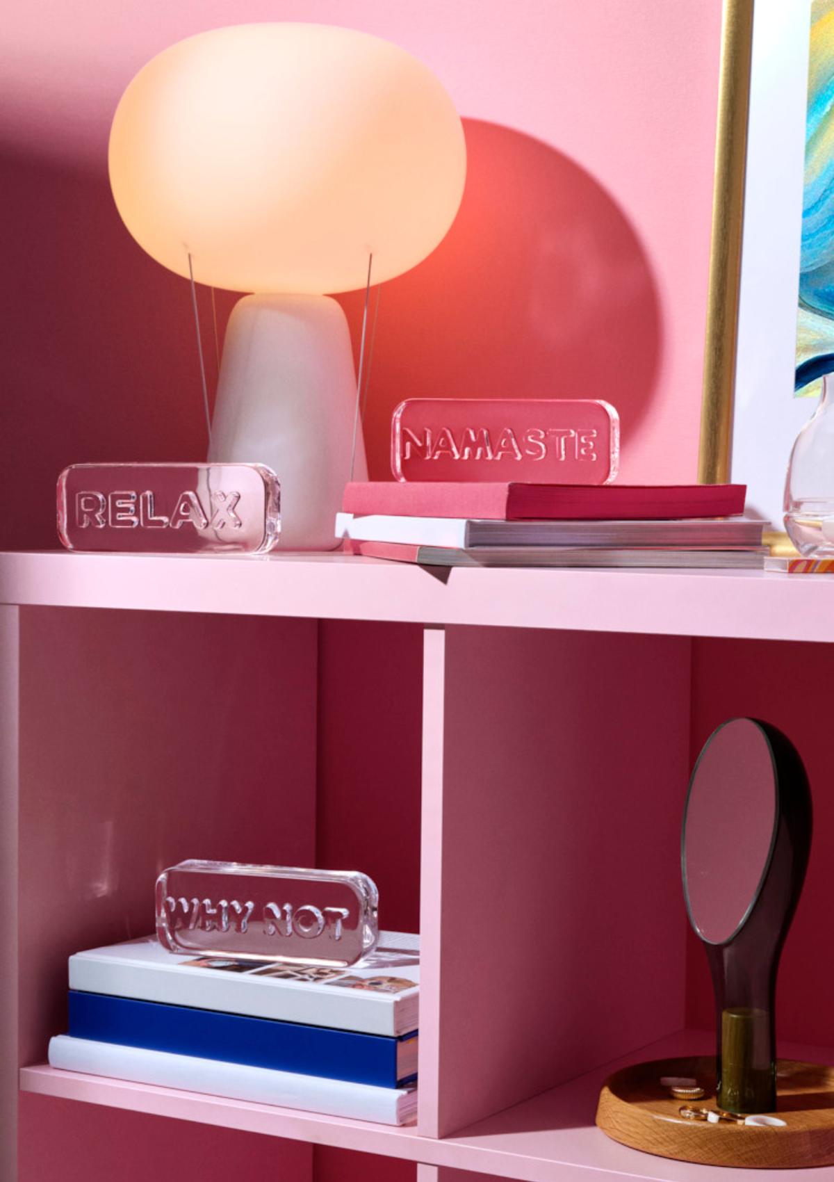 NUDE Paroles Relax, Namaste and Why Not paperweights, a Blow Lamp and Moon Mirror styled on a pink shelf against a deep berry wall.