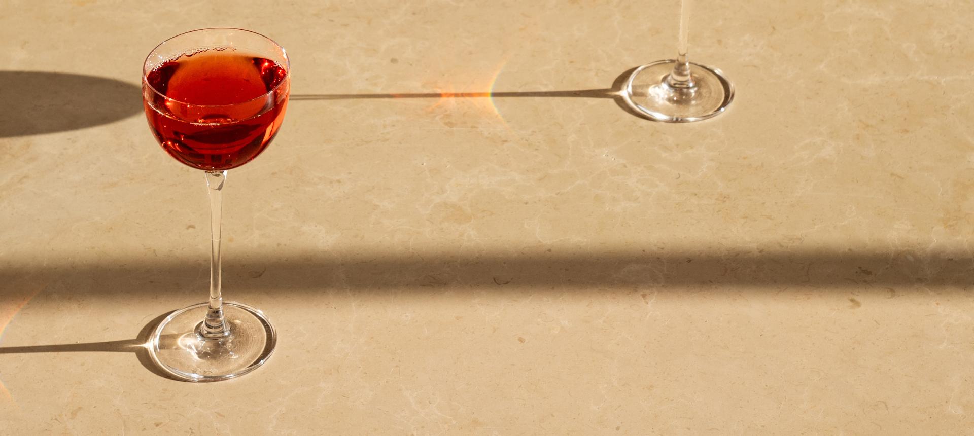 A Savage Pony glass filled with red cocktail sits on a warm beige stone surface, casting a long shadow. A second wine glass is partially visible in the upper right, and a bright light flare crosses the center of the frame.