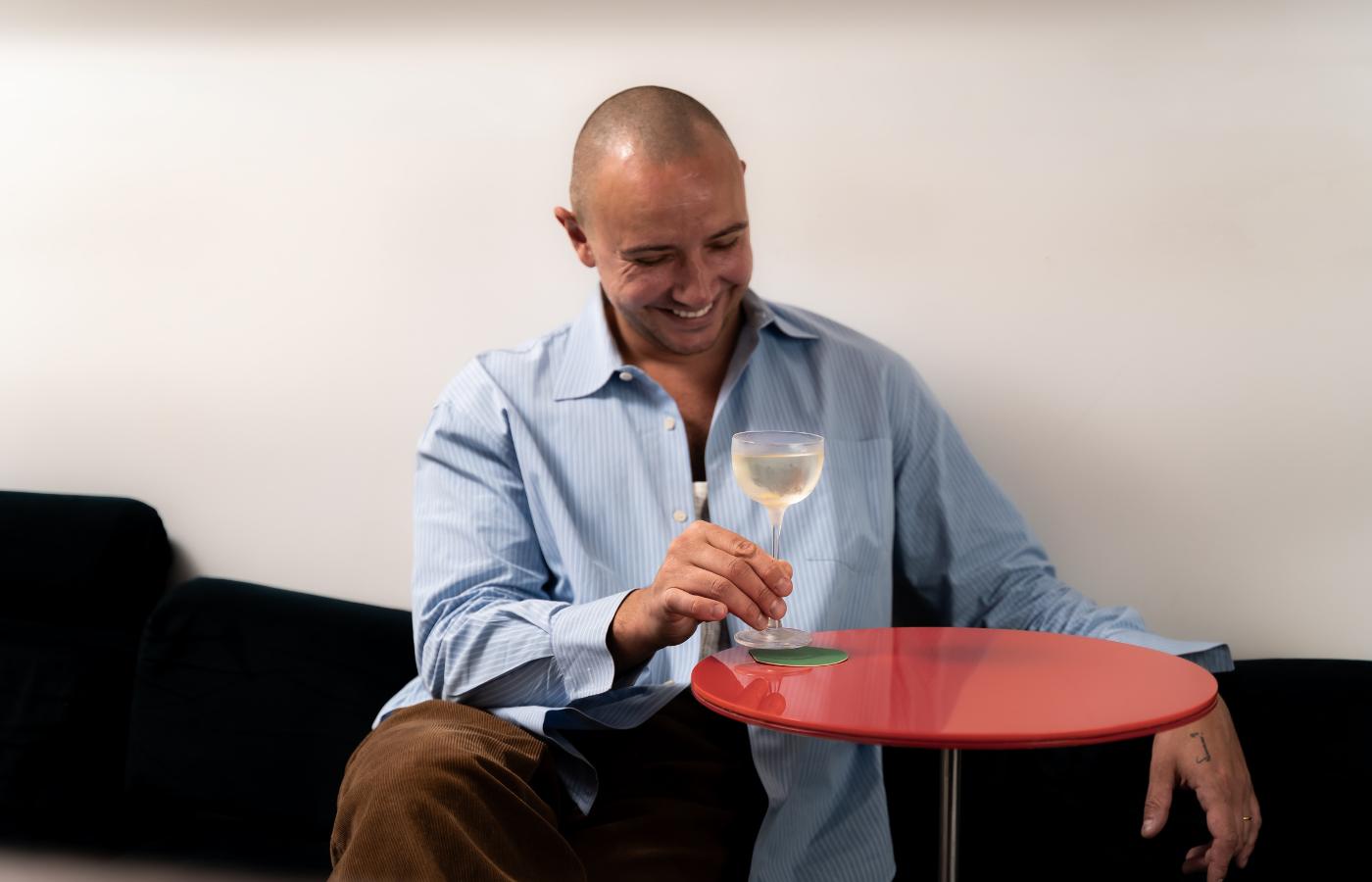 Rémy Savage seated with a cocktail in the NUDE Savage Pony Glass on a red side table