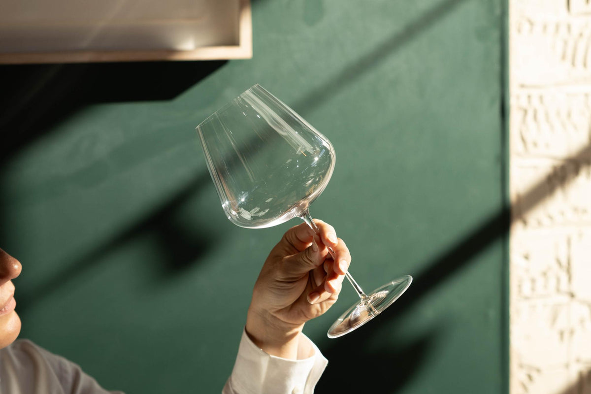 Honey Spencer tilting an empty NUDE Stem Zero Elegant Medium Red Wine Glass to the light against a green wall