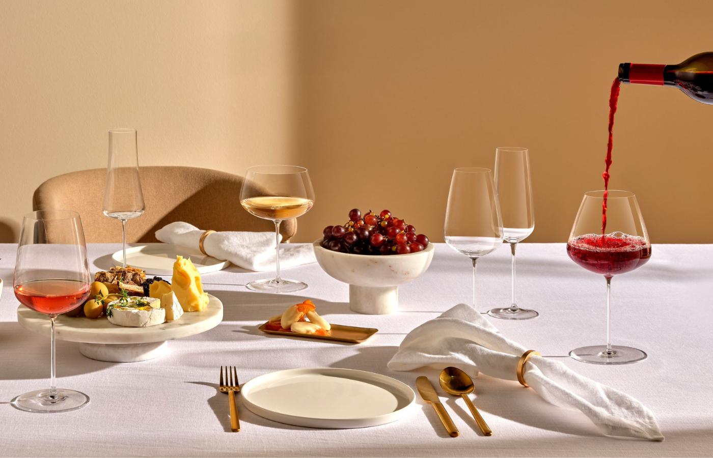 NUDE Stem Zero Vertigo wine glasses with red wine being poured, styled with white wine, grapes and gold cutlery on an elegant table setting.