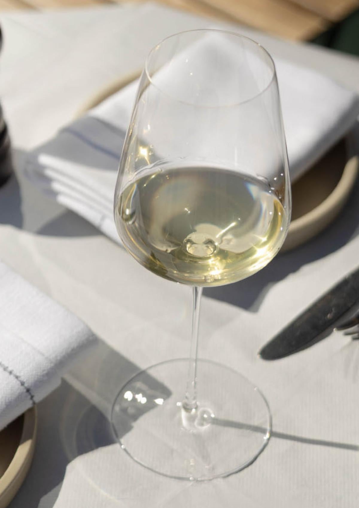 A NUDE Stem Zero Vertigo Delicate White Wine Glass filled with white wine on a sunlit linen table setting