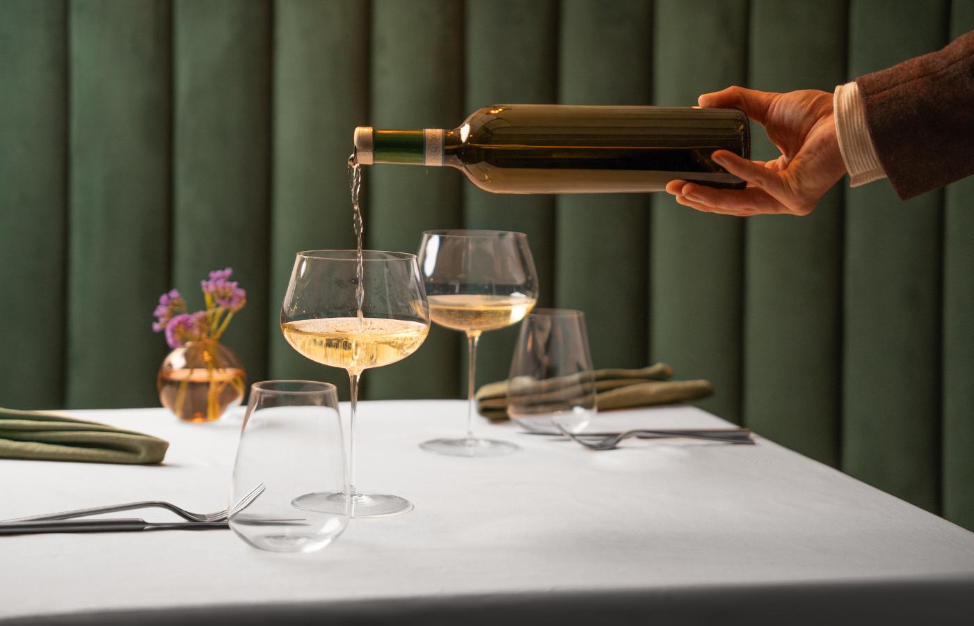 NUDE Stem Zero Vertigo Full Bodied White Wine Glass with white wine being poured in an elegant restaurant setting with green velvet walls