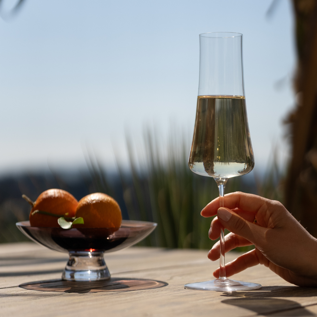 NUDE Stem Zero Volcano champagne glass held by hand on a sunlit outdoor terrace with citrus fruit bowl and open sky